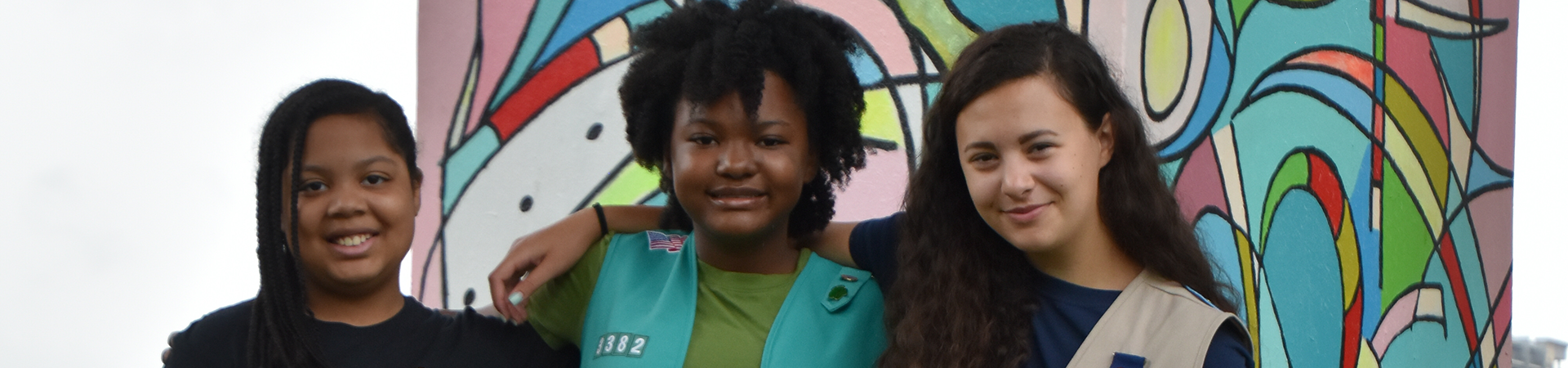  group of elementary school girl scouts outside next to graffiti wall talking - girl on left holds clip board  